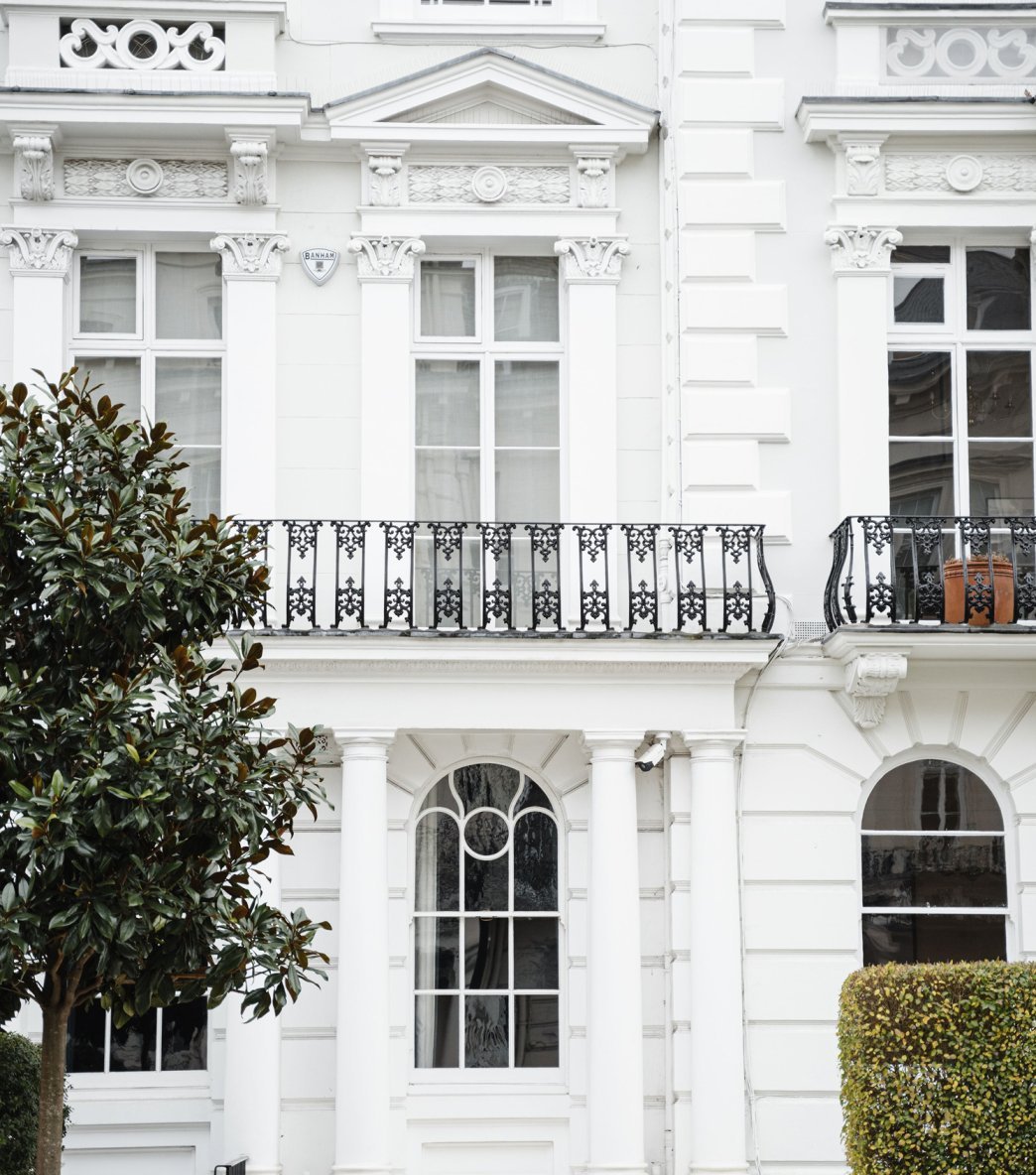 White multi-story building with ornate architectural details, black wrought iron balcony railing, large windows, enclosed by green shrubbery and trees.