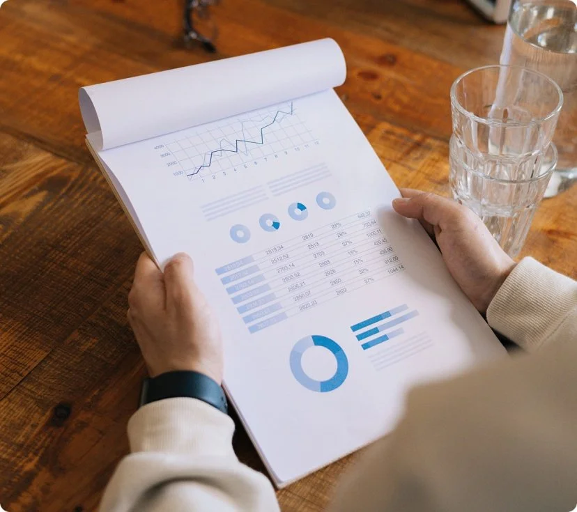 A person is reviewing a printed report with various charts and graphs, including line, pie, and bar graphs, on a wooden table with three glasses of water nearby.