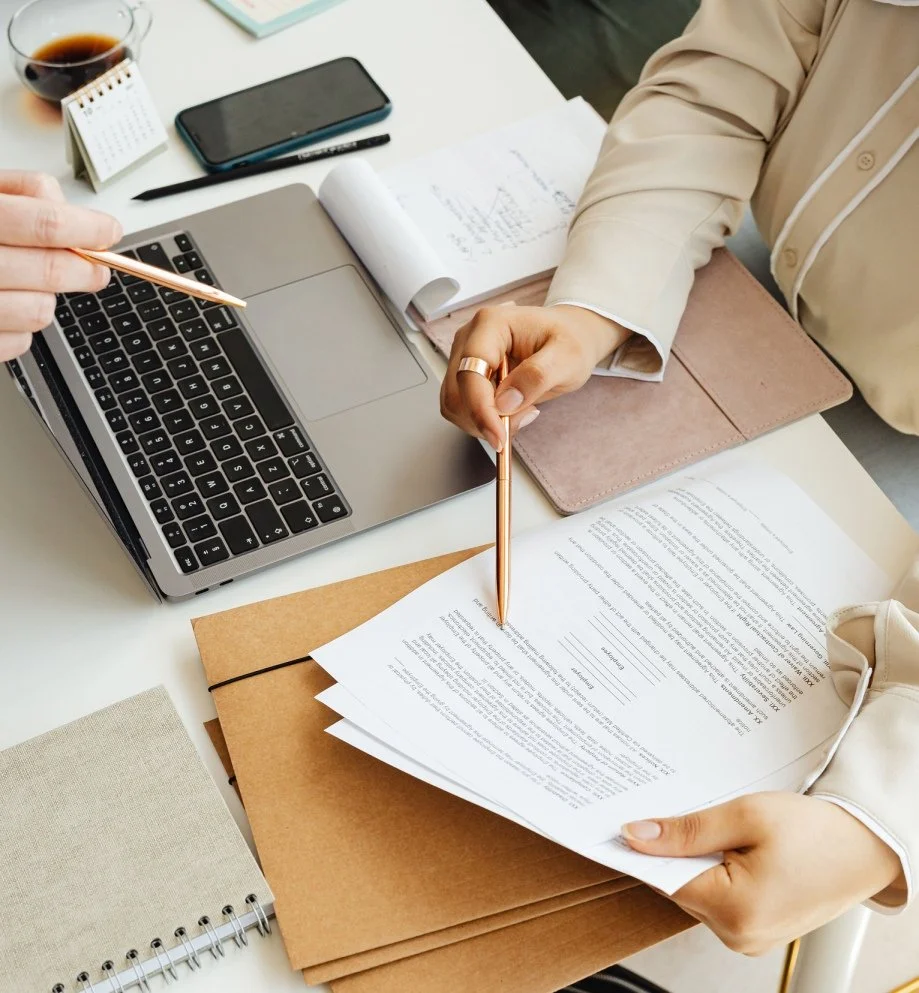 A person is reviewing documents at a desk with a laptop, a smartphone, a notebook, and a glass of red wine.