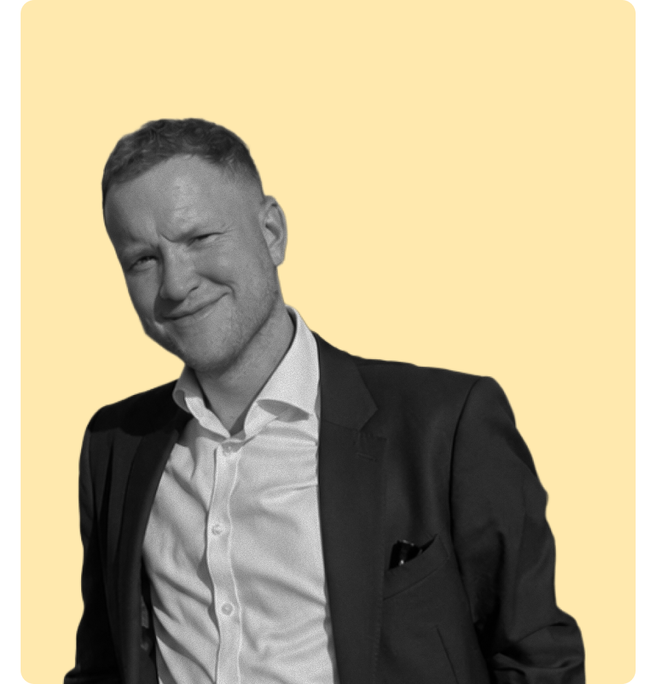 A man with short hair wearing a white shirt and dark suit smiling and winking, against a yellow background.