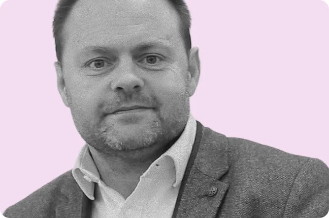 A portrait photograph of a man with short hair, a beard, wearing a white shirt and a grey blazer, against a light purple background.