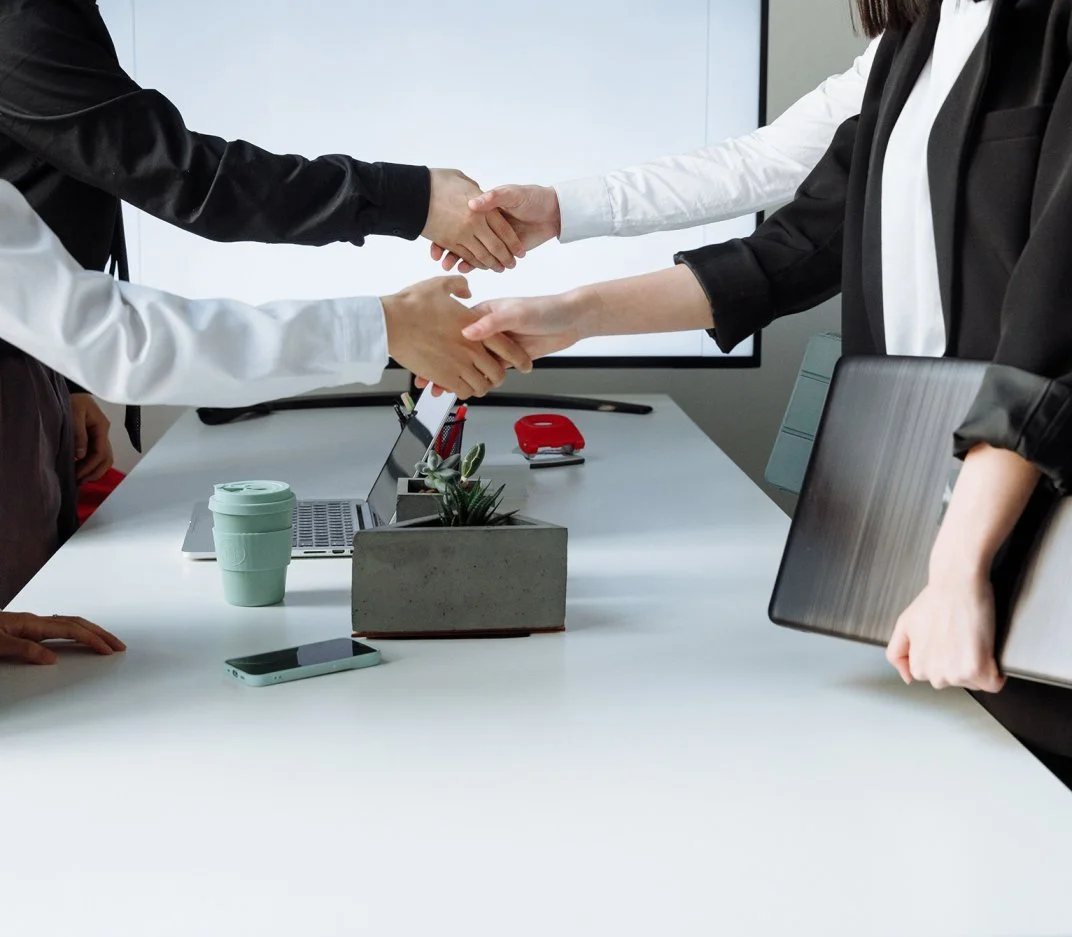 Two people shaking hands across a conference table with laptops, a phone, and office supplies on it.