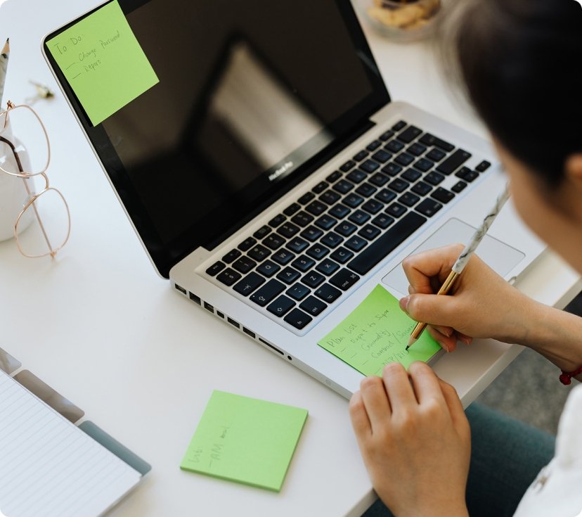 Person writing notes on green sticky notes at a laptop desk.