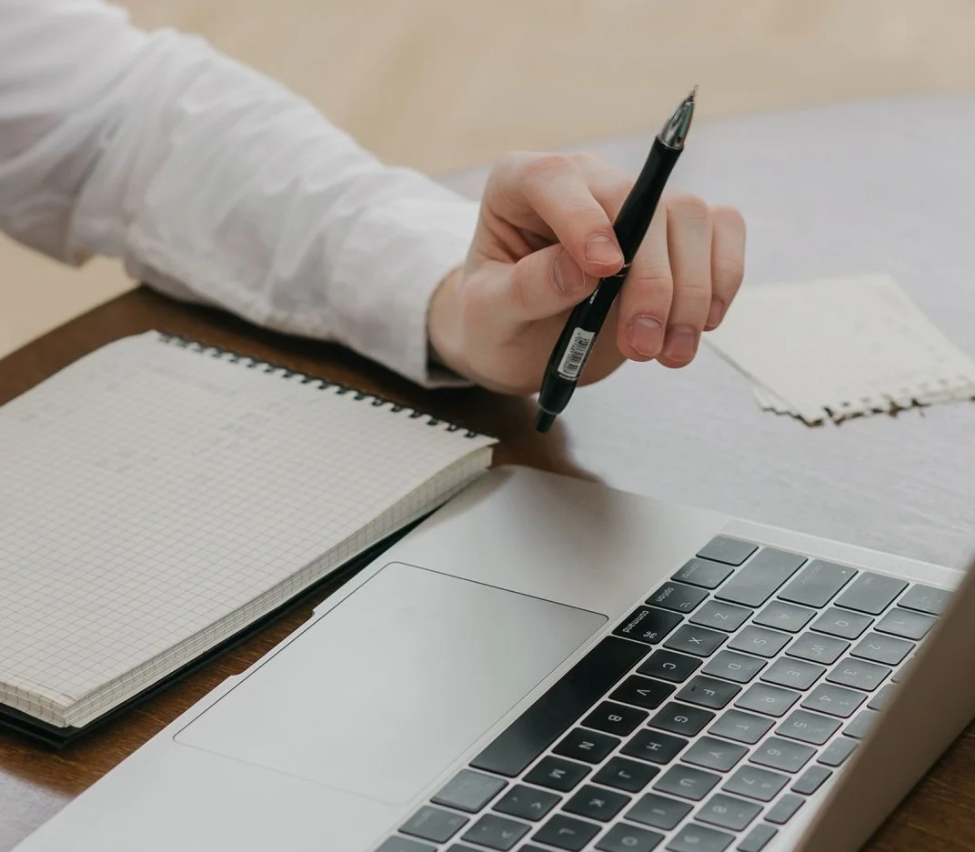 Person holding a black pen near a laptop keyboard, with a spiral notebook and small pieces of paper on a wooden table.