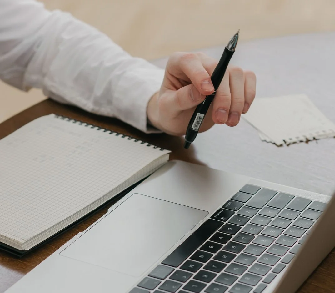 Person holding a black pen over a laptop with a grid notebook nearby.