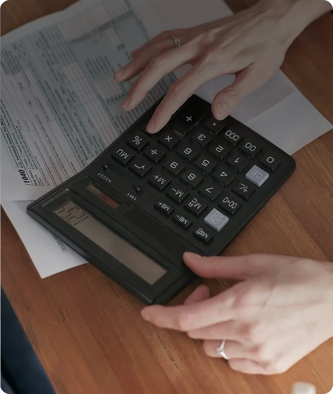 A person is using a calculator on a wooden table, with documents and papers underneath.