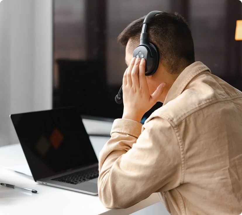 A man wearing a beige jacket and large black headphones sits at a desk with a laptop, with a pen nearby, in a modern workspace.
