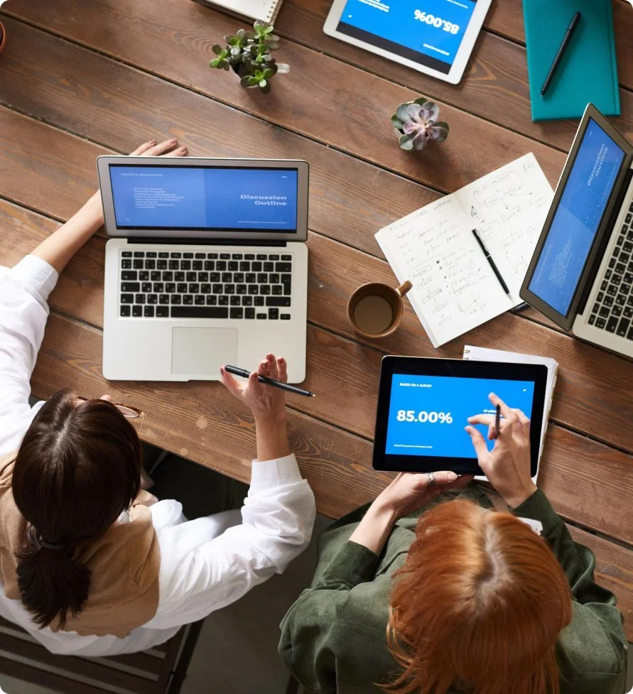 Two women sitting at a wooden table with laptops, tablets, notebooks, and a cup of coffee, engaged in a meeting or work session, with potted plants on the table.