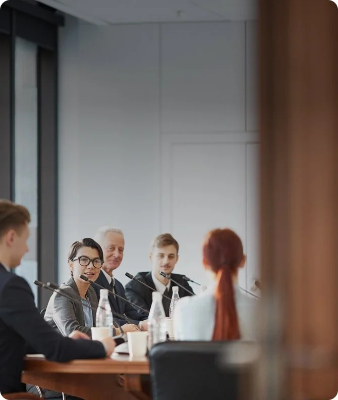 A group of professionals sitting at a conference table in a meeting room, engaged in discussion, with microphones and water bottles in front of them.