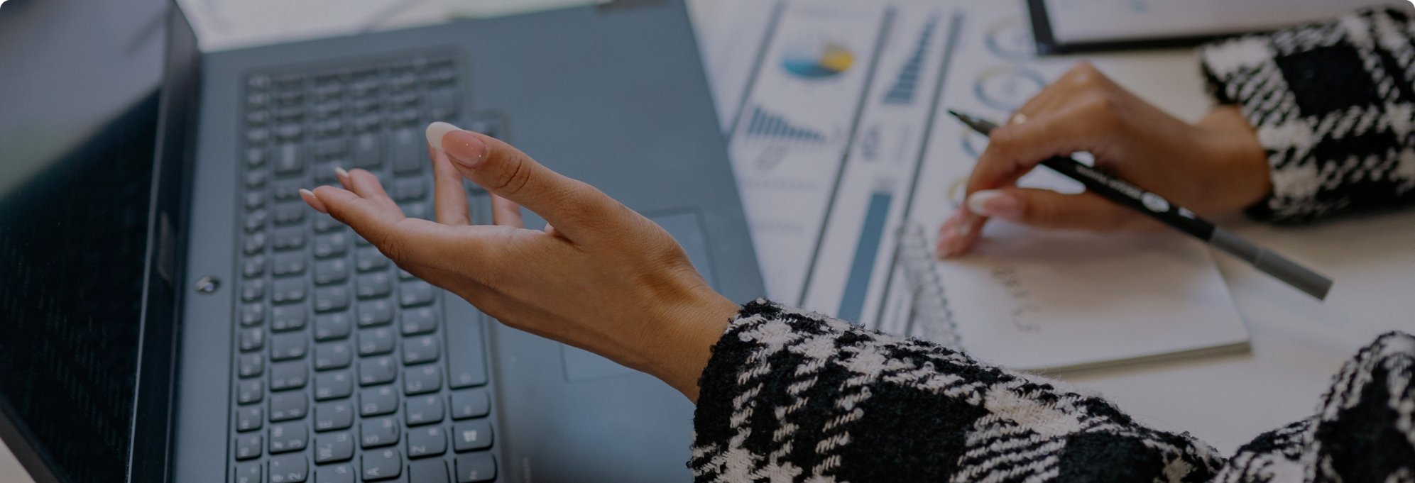A person in a black and white patterned sweater working on a laptop with documents and charts on the desk.