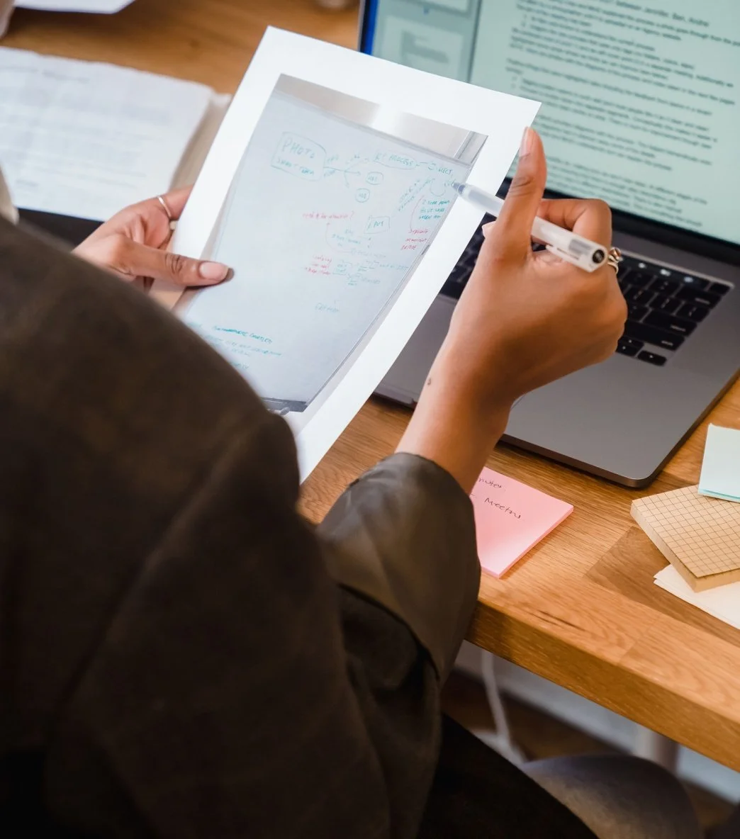 Person holding a photographic printout and writing on it with a pen at desk with open laptop, sticky notes, and papers.