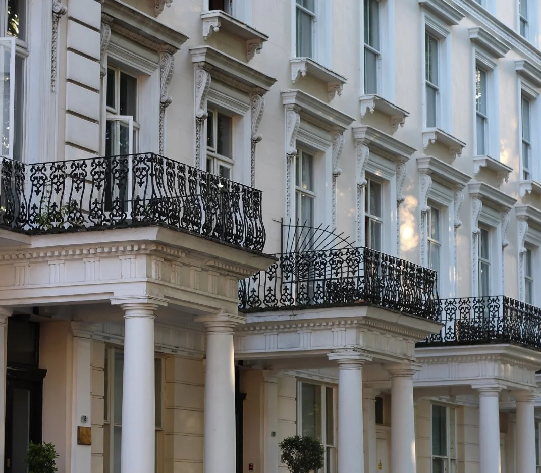 White multi-story building with ornate architectural details, black wrought iron balcony railings, and tall columns supporting the porch, featuring multiple windows with decorative moldings.