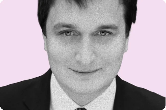 Black and white portrait of a man wearing a suit and tie, with a slight smile, against a light pink background.