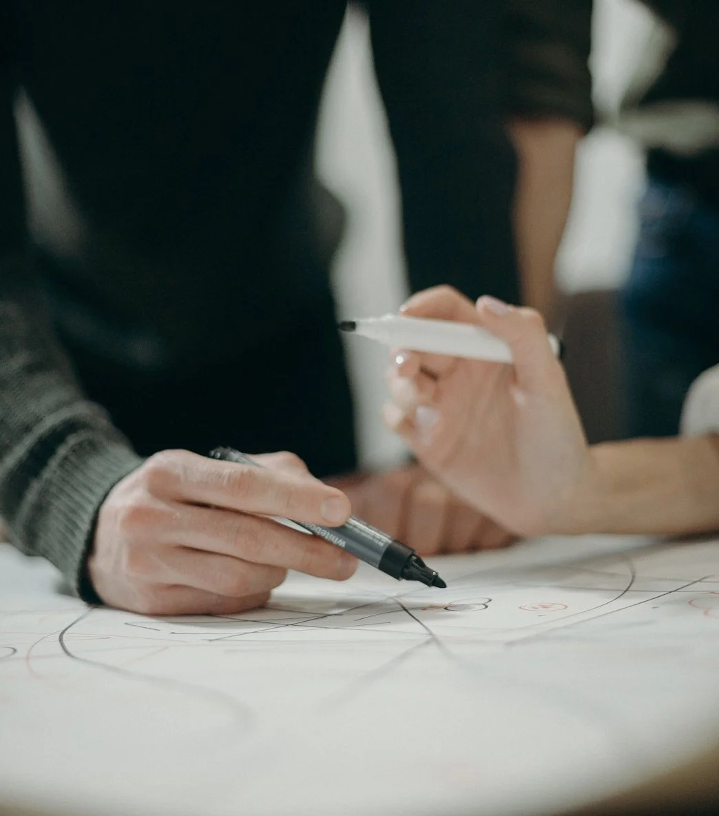 Close-up of a person's hands sketching with a black marker and holding a white marker, on a large sheet of paper with sketches or diagrams.