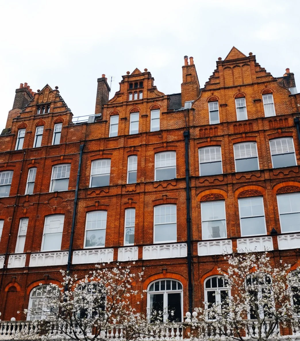 Red brick Victorian-style building with ornate architectural details and tall chimneys, overlooking flowering trees.