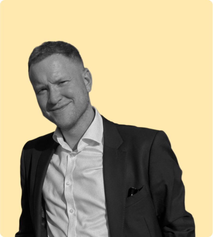 Black and white photo of a man in a suit smiling and looking into the camera, standing against a light yellow background.