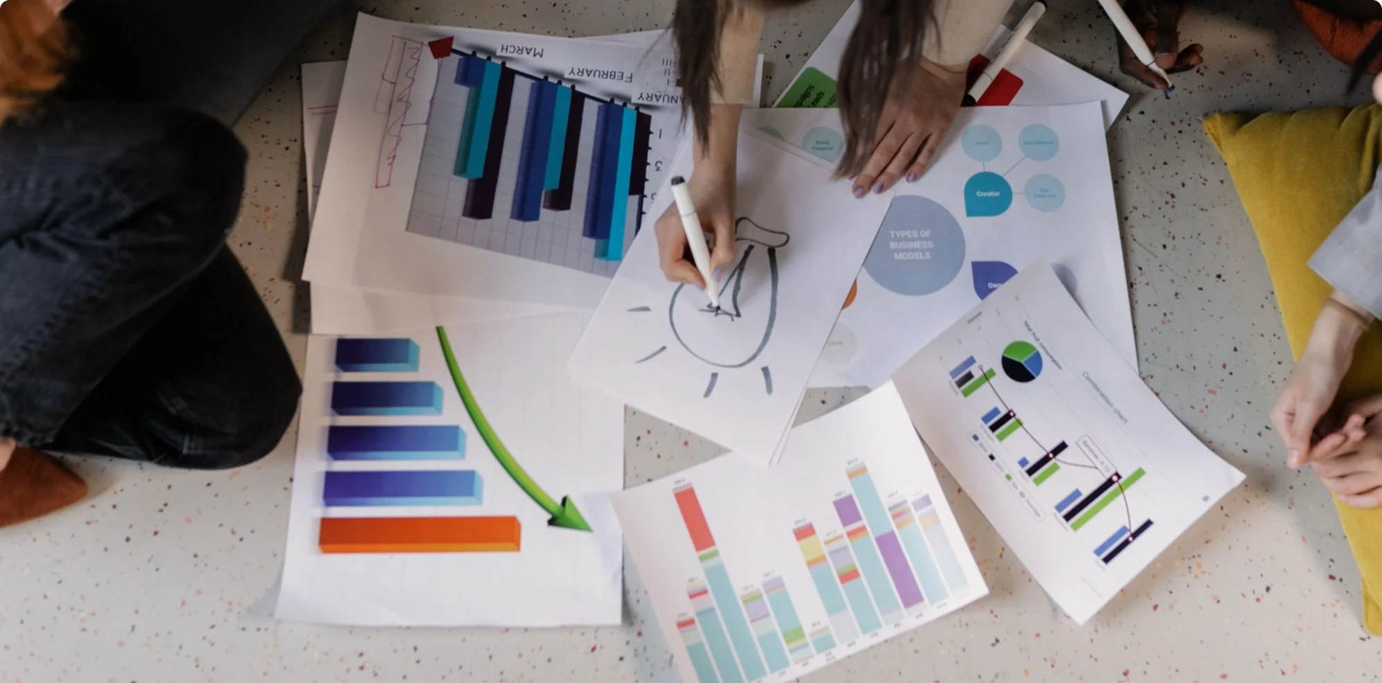 Overhead view of a group of people working on various business charts and graphs spread out on a speckled floor, with one person drawing on a paper with a marker.