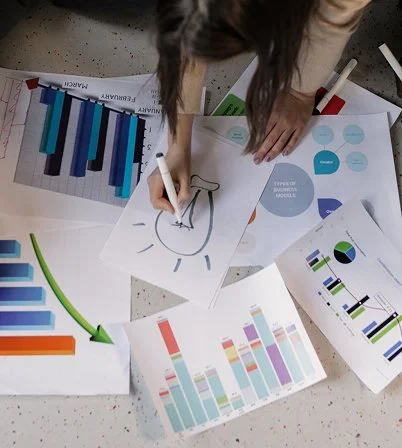 Person using a white marker on paper with a drawing of a hammer and rays, surrounded by various colorful charts and graphs on a table.