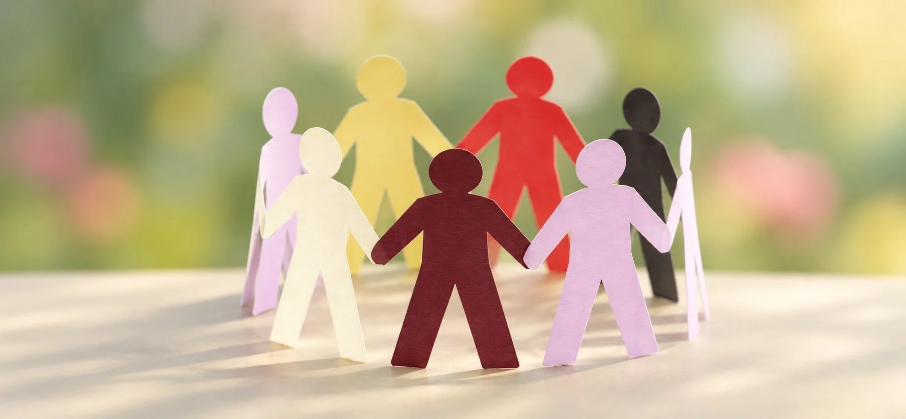 Colorful paper cutout figures hold hands in a circle against a blurred natural background.