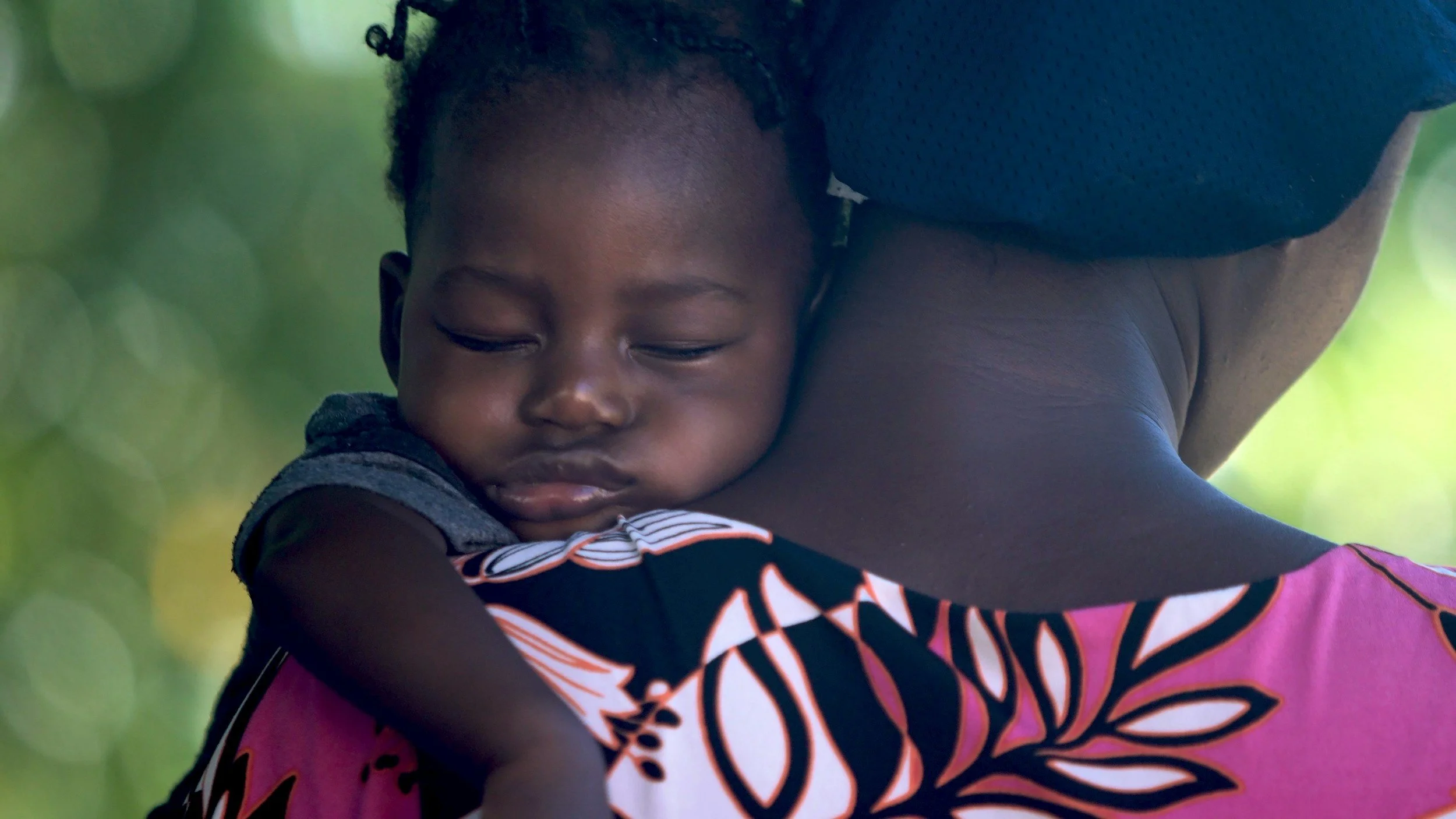 A young child with dark skin and short curly hair sleeping on an adult's shoulder outdoors with a blurred green background.
