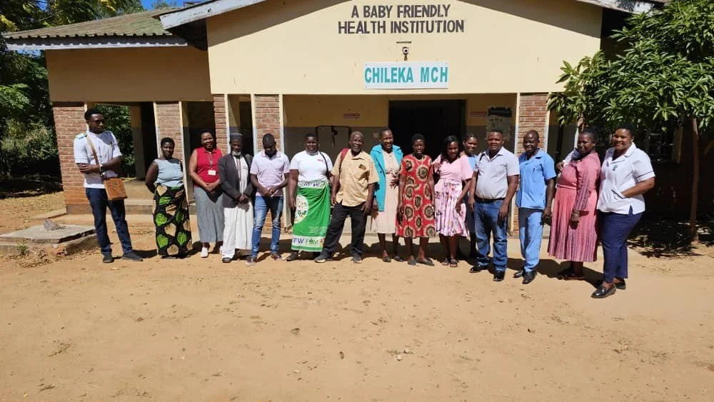Group of people standing in front of a building with a sign that reads 'A Baby Friendly Health Institution CHILEKA MCH.' The group includes men and women, some in traditional clothing, standing on a dirt surface.