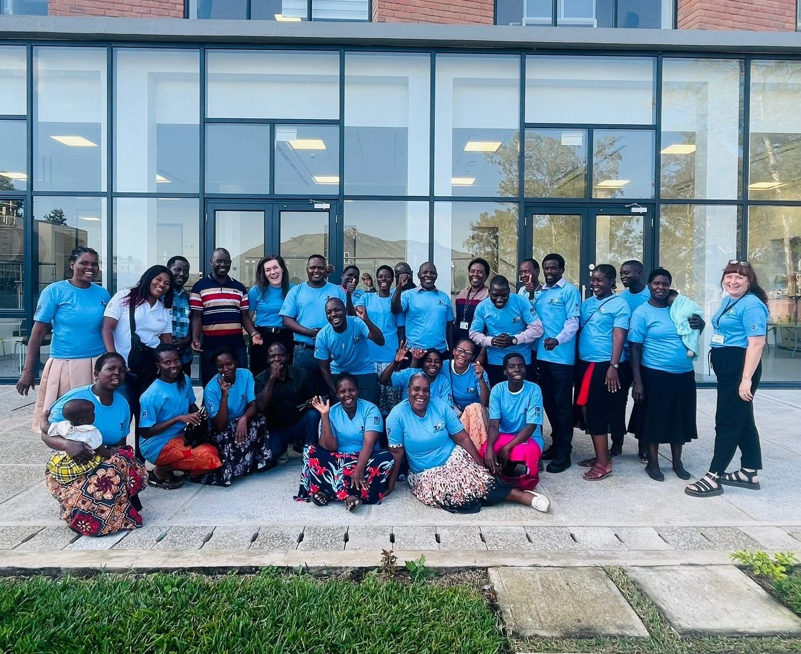 A large group of people, most wearing blue shirts, are gathered outside a modern glass building, smiling and posing for a group photo.