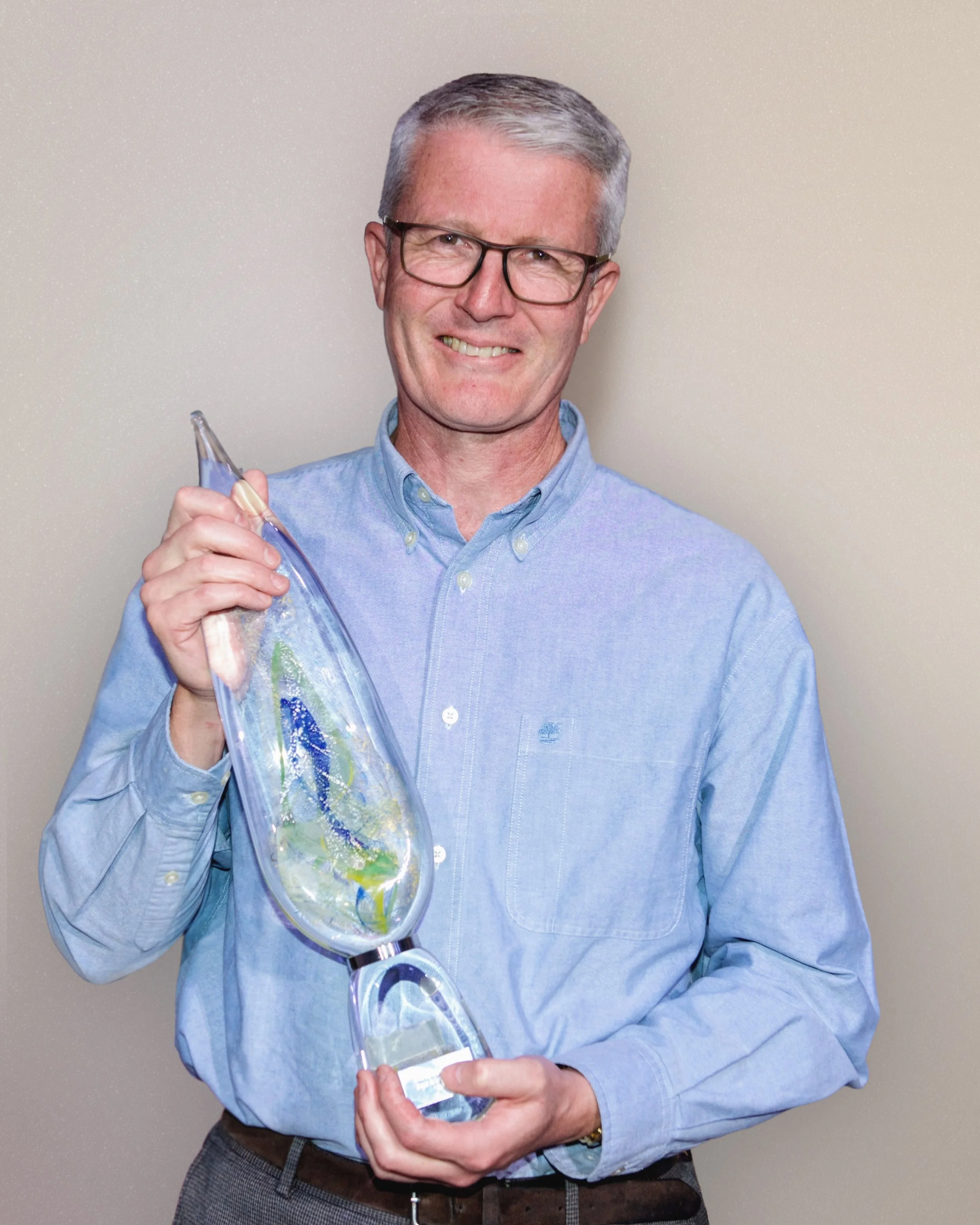 A man with gray hair and glasses smiling while holding a glass art piece that resembles a fish.