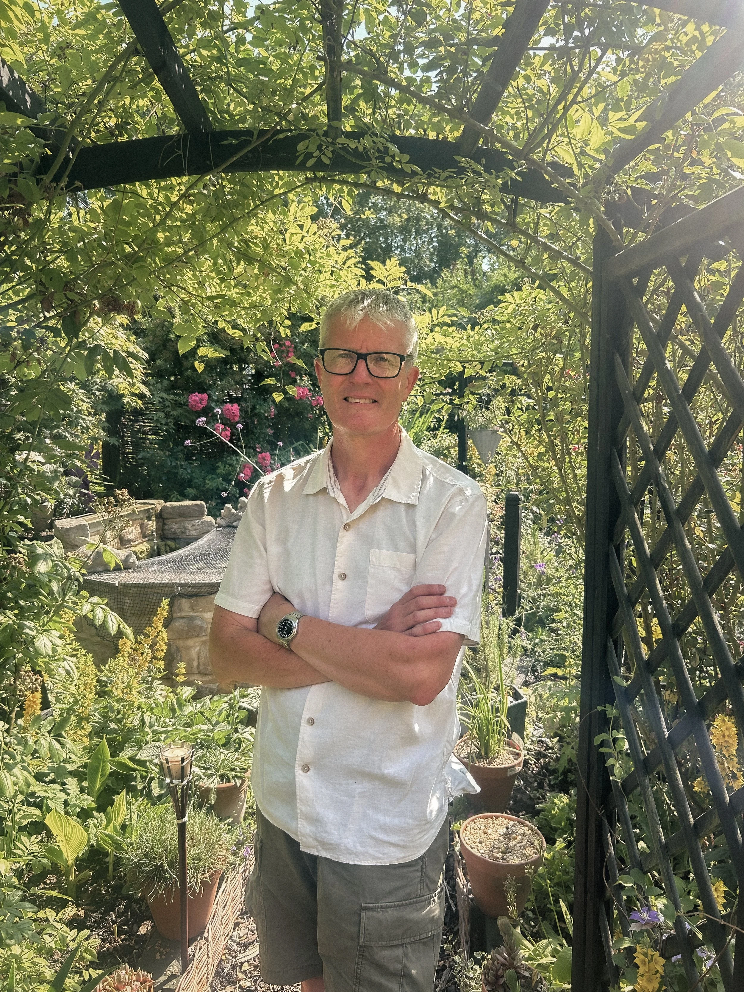 A man with glasses and a white button-up shirt standing in a lush garden with various plants and pink flowers, under a black archway structure.
