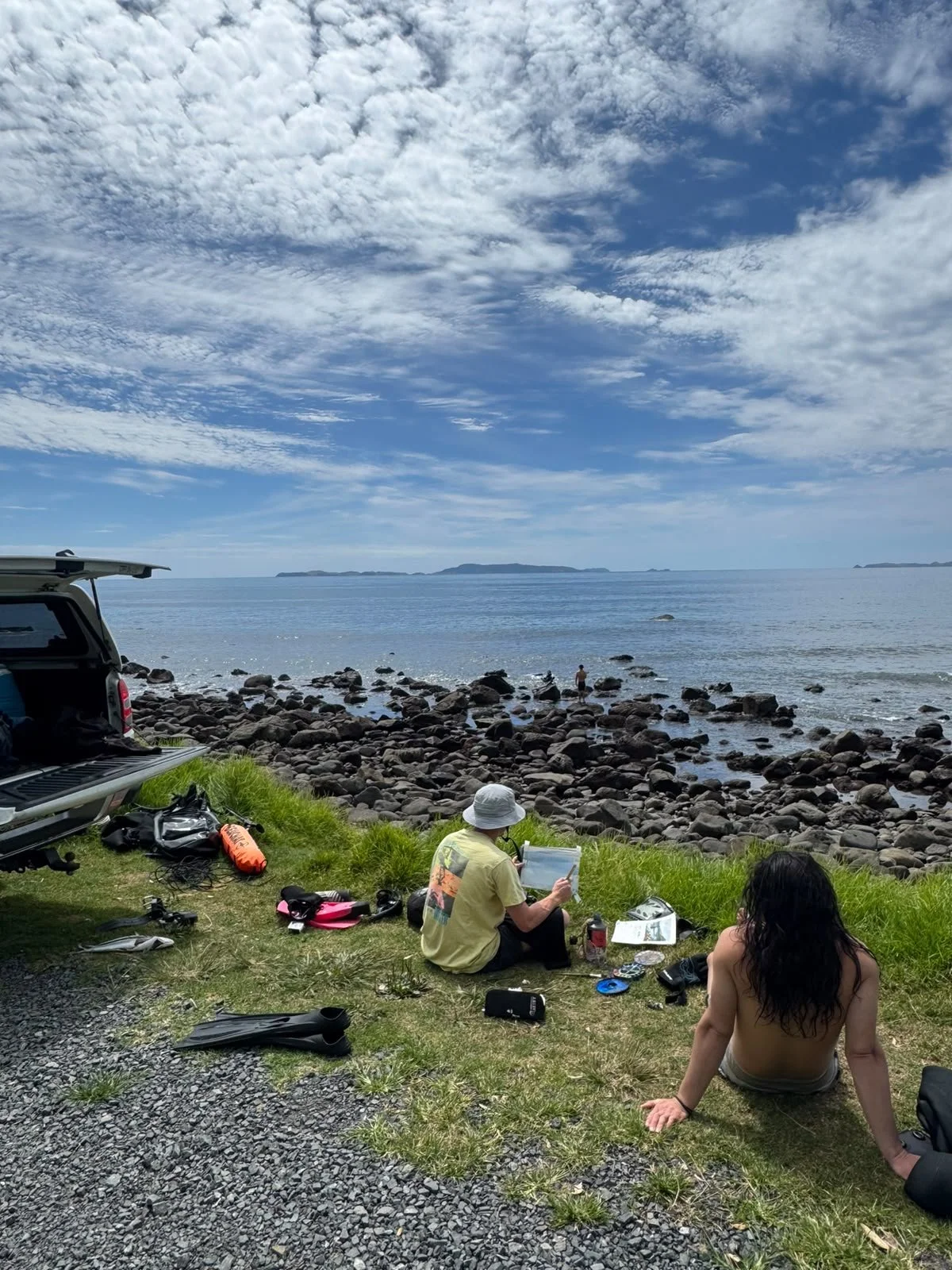 People sitting on grassy area near rocky shoreline, ocean view with clouds and distant islands, open car trunk with gear nearby.