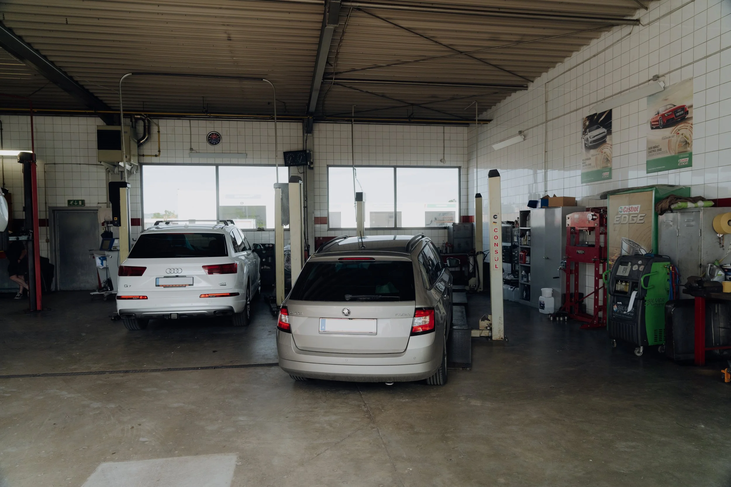 Innenansicht einer Autowerkstatt mit mehreren geparkten Fahrzeugen, Werkzeugen und Poster an der Wand.