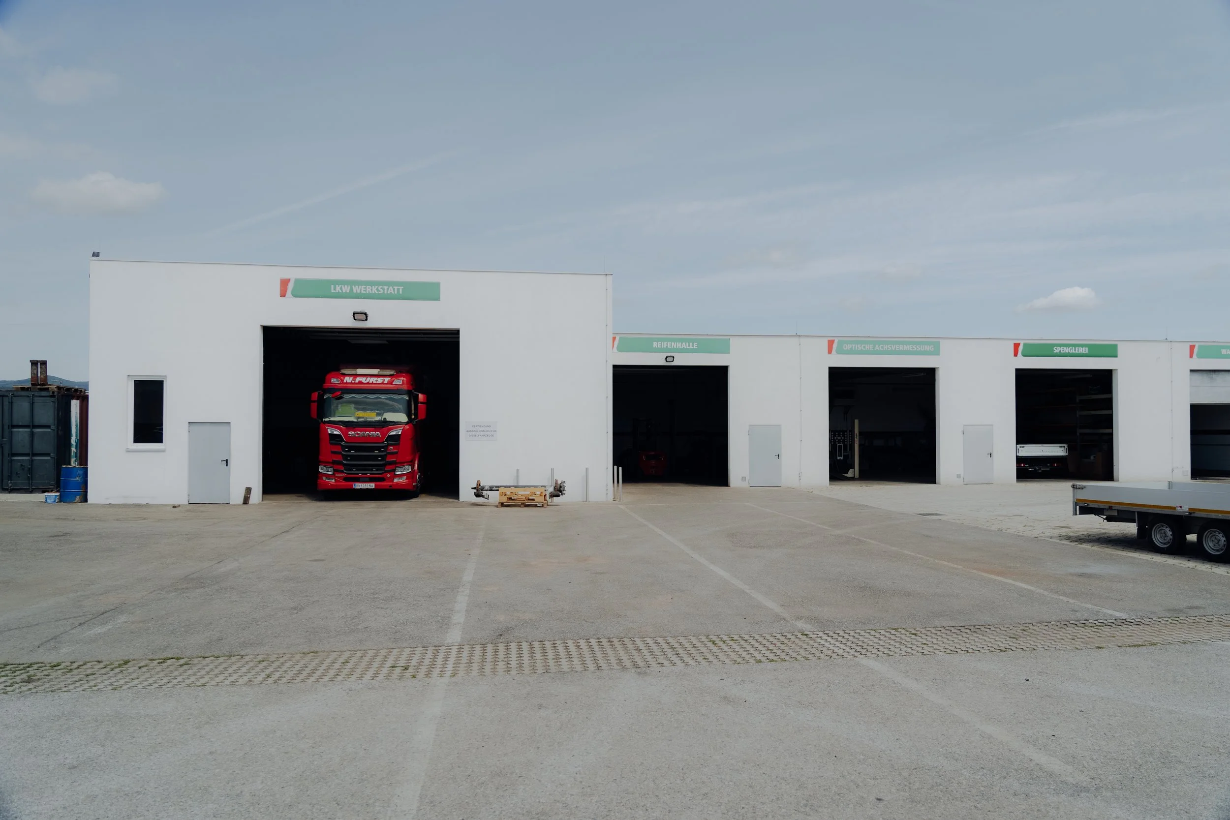 Ein weißes Werkstattgebäude mit mehreren Garagen, in einer Garage steht ein roter Lastwagen. Über den Garagen sind grüne Schilder mit deutschen Beschriftungen, z.B. 'LKW Werkstatt', 'Reifenhalle', 'Optische Achsvermessung' und 'Spenglerei'. Der Himmel ist teilweise bewölkt und der Boden ist gepflastert.