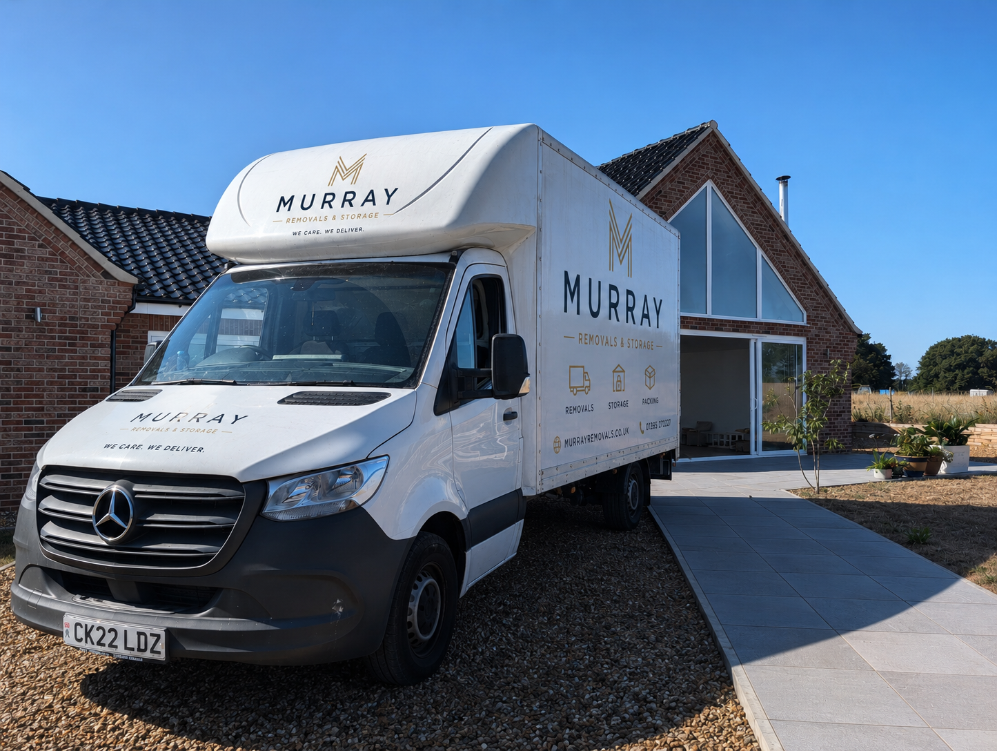 A white removal and storage truck labeled 'MURRAY REMOVALS & STORAGE' parked in front of a modern brick house with large glass windows under a blue sky.