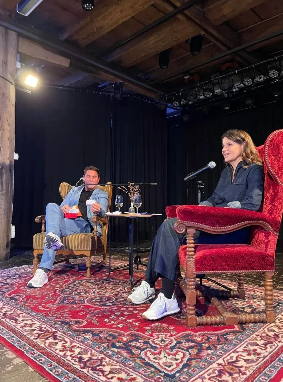 Two people sitting in chairs on a stage with a microphone, glasses, and a small table between them, engaging in a conversation or interview, with a dark curtain as the backdrop.