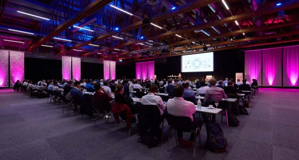 Conference room filled with attendees listening to a speaker on stage, with a large screen displaying a presentation, purple lighting, and high wooden ceilings.
