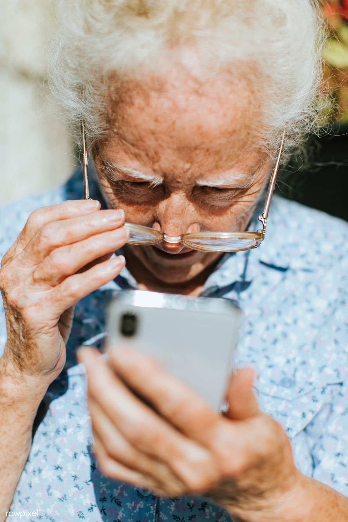 An elderly woman with white hair and glasses looking at her phone, with her hand adjusting her glasses and her other hand holding the phone.