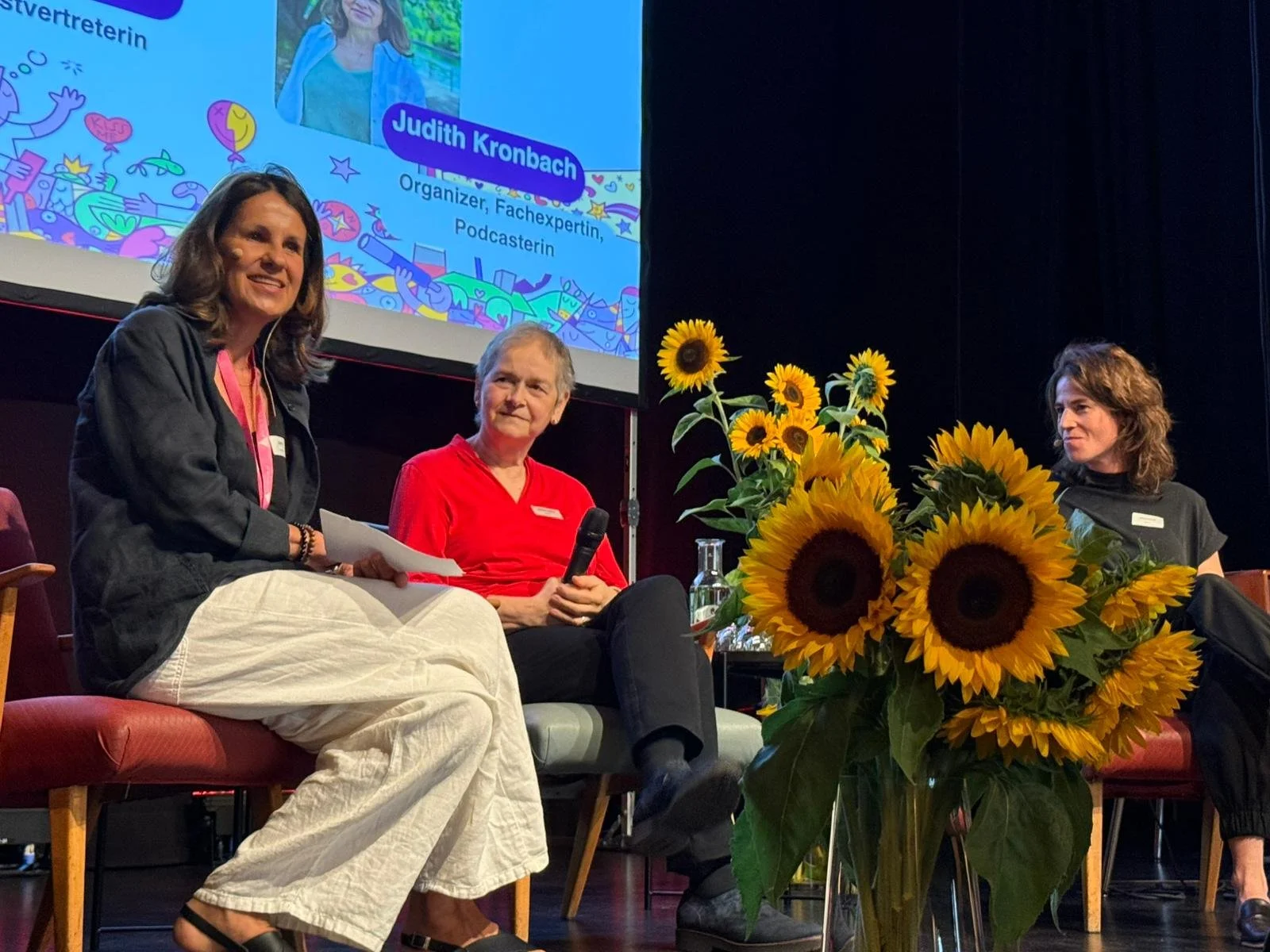 A panel discussion featuring three women sitting on a stage with a large bouquet of sunflowers in the foreground. One woman is speaking into a microphone, while the other two listen. A large screen behind them displays a colorful illustration, a photo of Judith Kronbach, and text indicating her as an organizer, Fachexpertin, and podcaster.