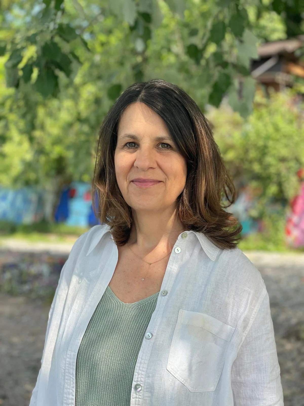 A picture of Judith Kronbach. A woman with shoulder-length dark brown hair with lighter brown highlights, wearing a white button-up shirt over a green top, stands outdoors in front of green leafy trees, smiling softly.