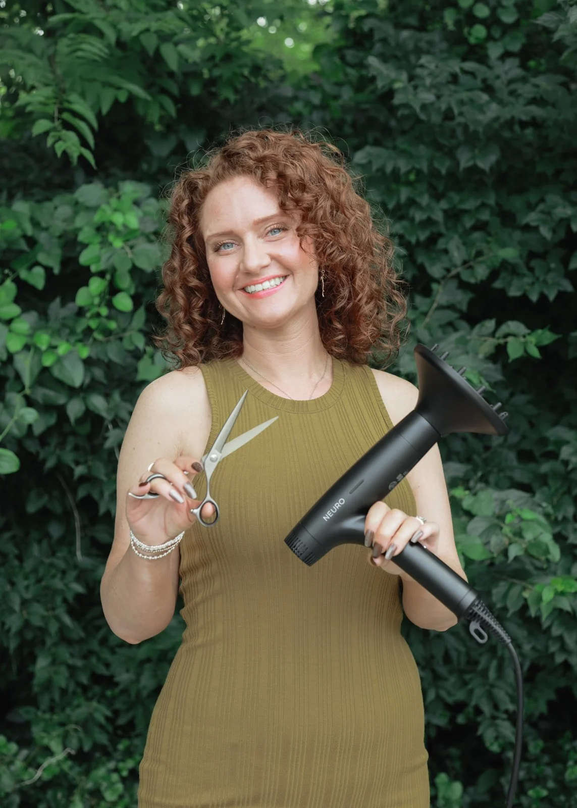 A woman with curly red hair smiling outdoors, holding hair-cutting scissors in one hand and a hairdryer in the other, with green leafy background.