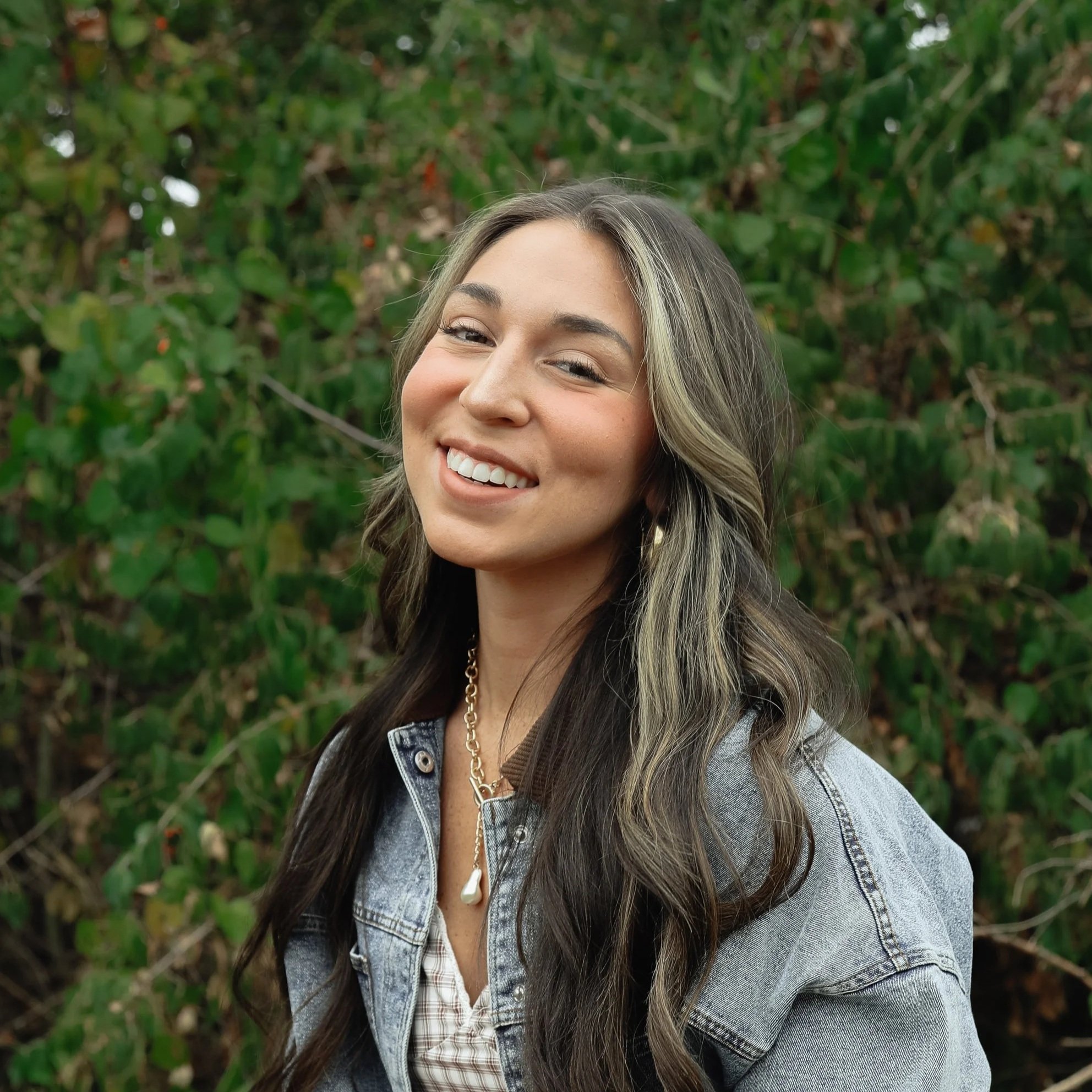A smiling woman with long hair and a denim jacket standing outdoors with green bushes in the background.