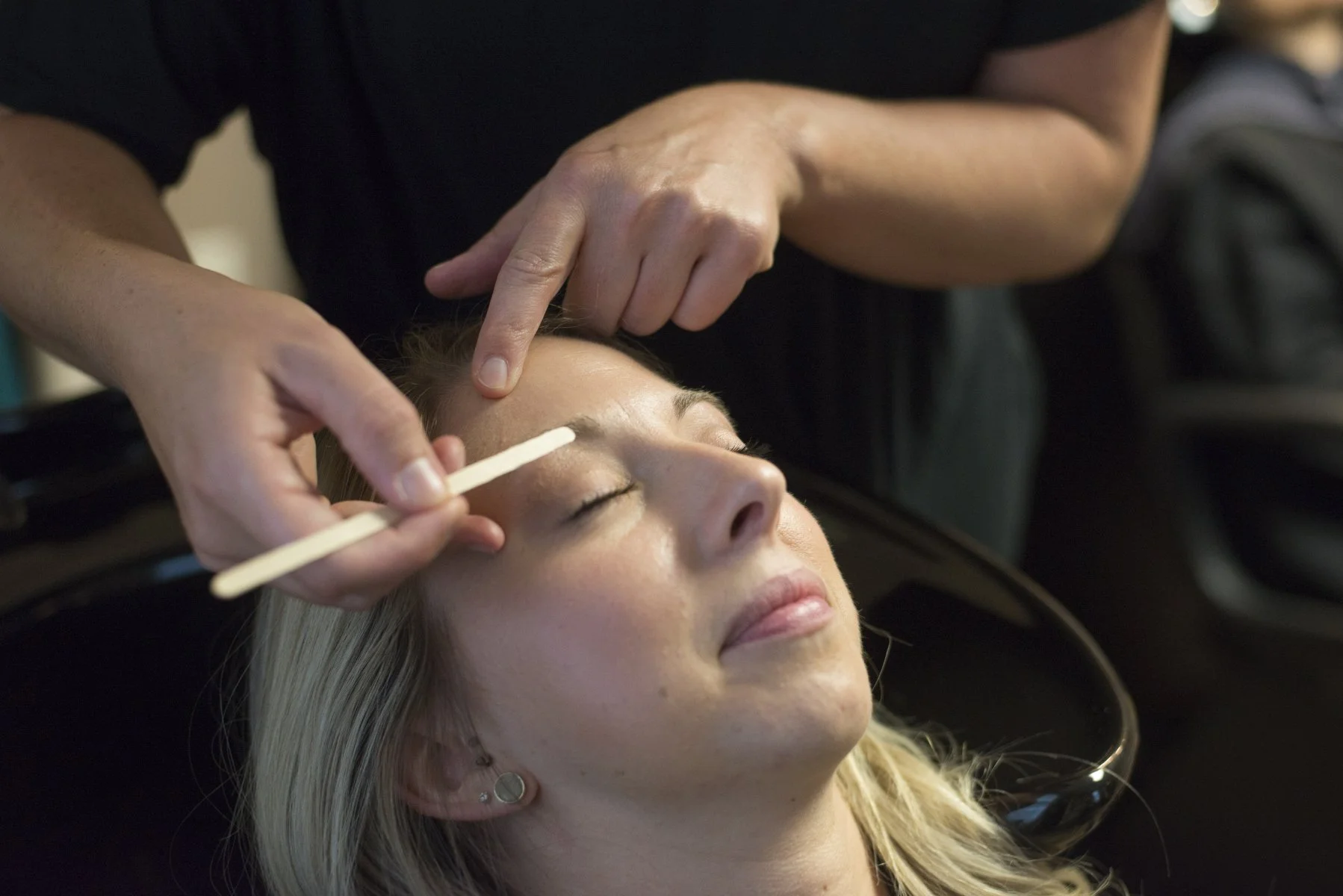 A woman with blonde hair receiving an eyebrow treatment at a salon, lying back with eyes closed, while a technician uses a small wooden stick near her eyebrow.