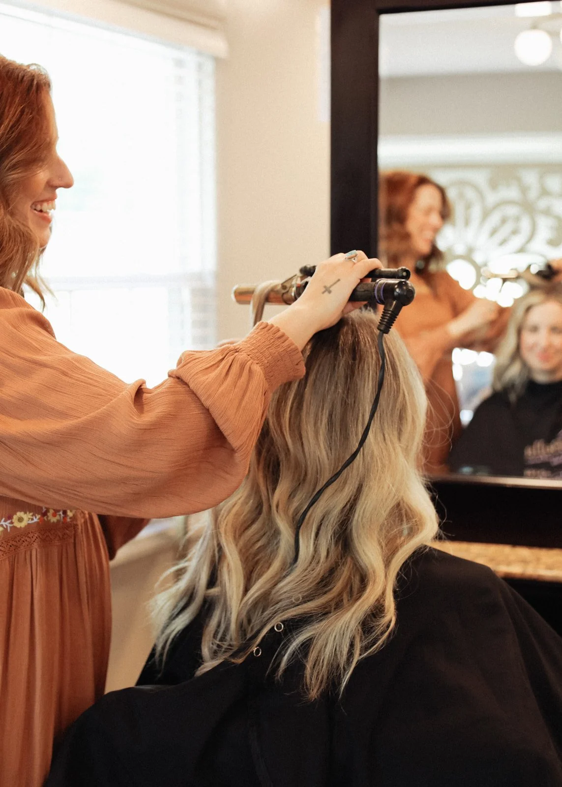 A hairstylist is curling a blonde woman's hair with a curling iron in a salon, seen through a mirror.