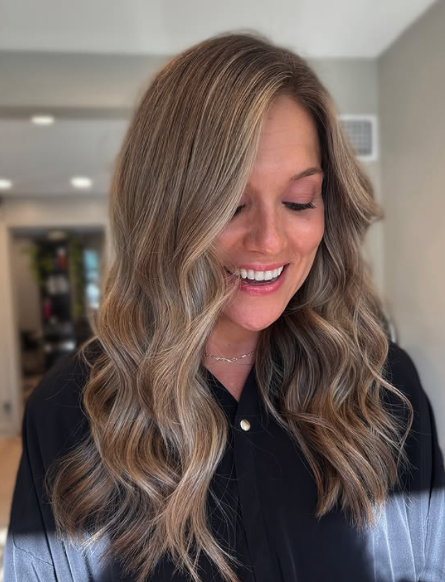A woman with long, wavy blonde hair smiling and looking down indoors with a blurred background.