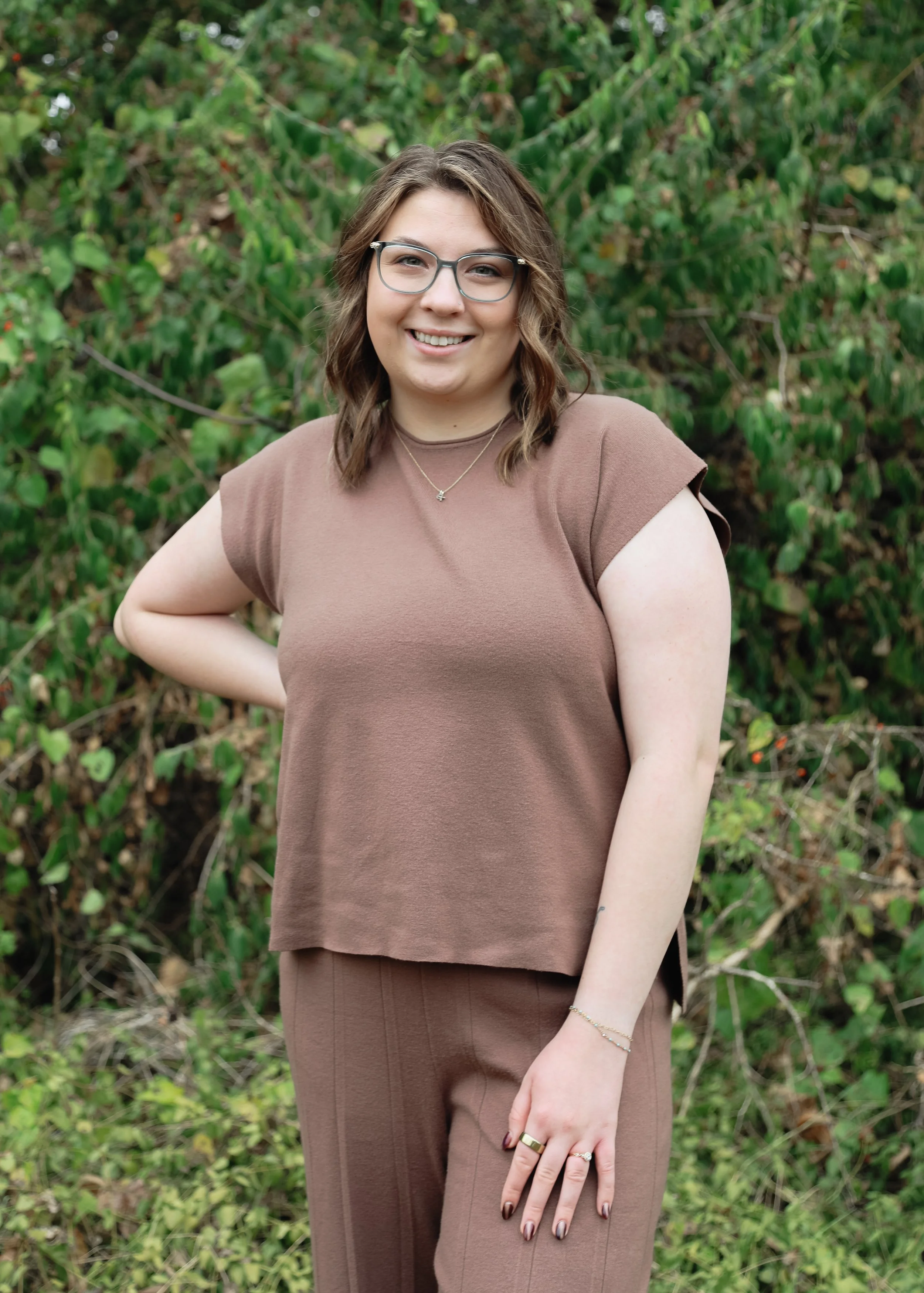 A smiling woman with glasses and wavy brown hair, wearing a brown top and matching pants, standing outdoors with a background of green foliage.