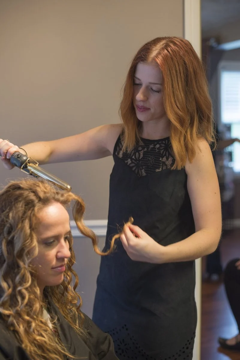 A hairstylist styles a woman's curly hair with a curling iron in a salon.