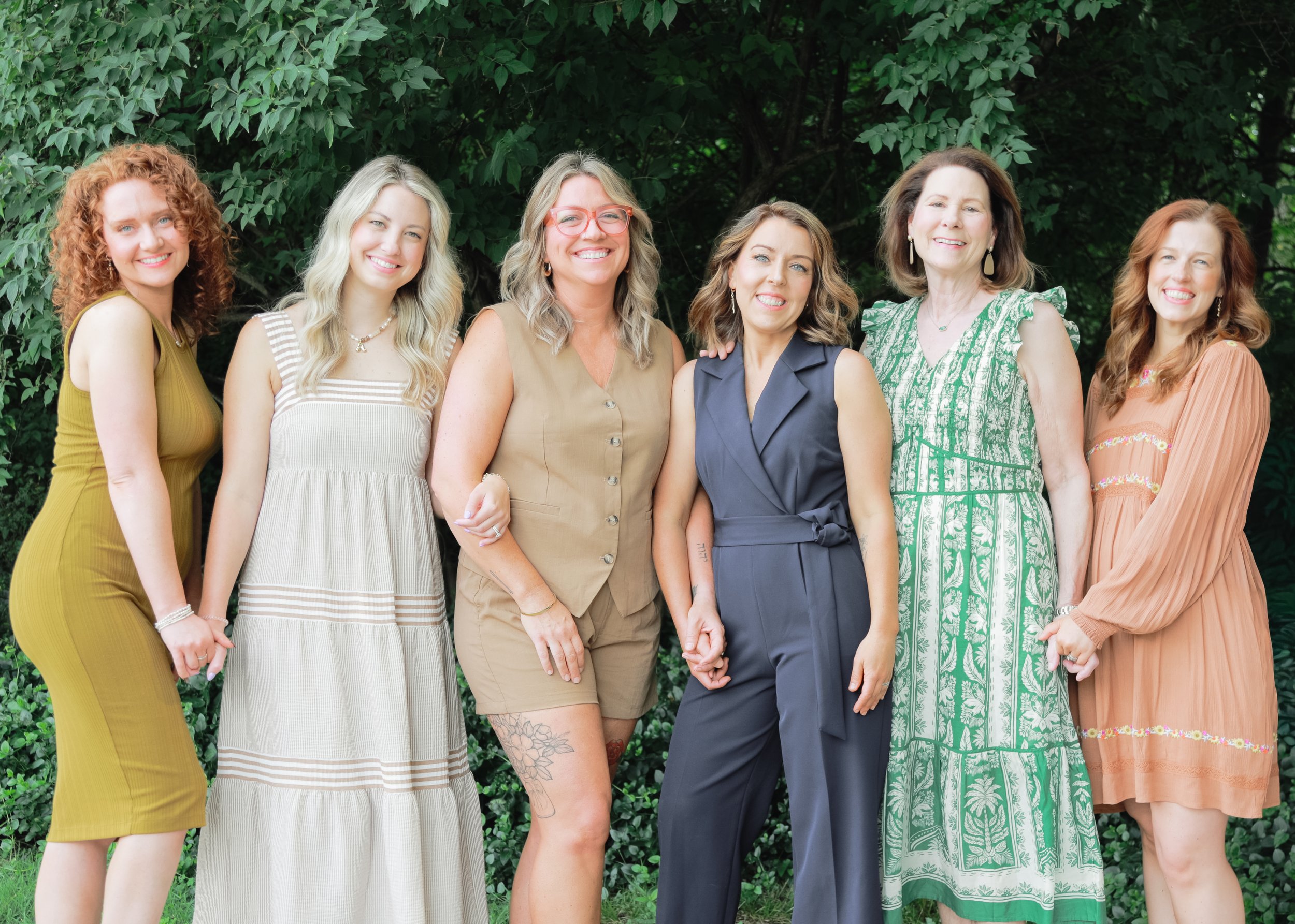 Group of seven women standing outdoors in front of green foliage, smiling at the camera.