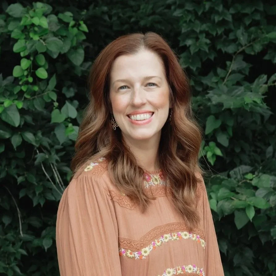 A woman with long wavy brown hair smiling, wearing earrings and a brown embroidered blouse, standing outdoors in front of green leafy bushes.