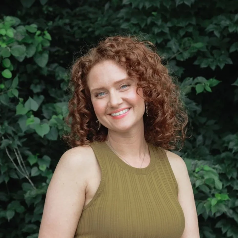A woman with curly red hair smiling outdoors with green foliage in the background.