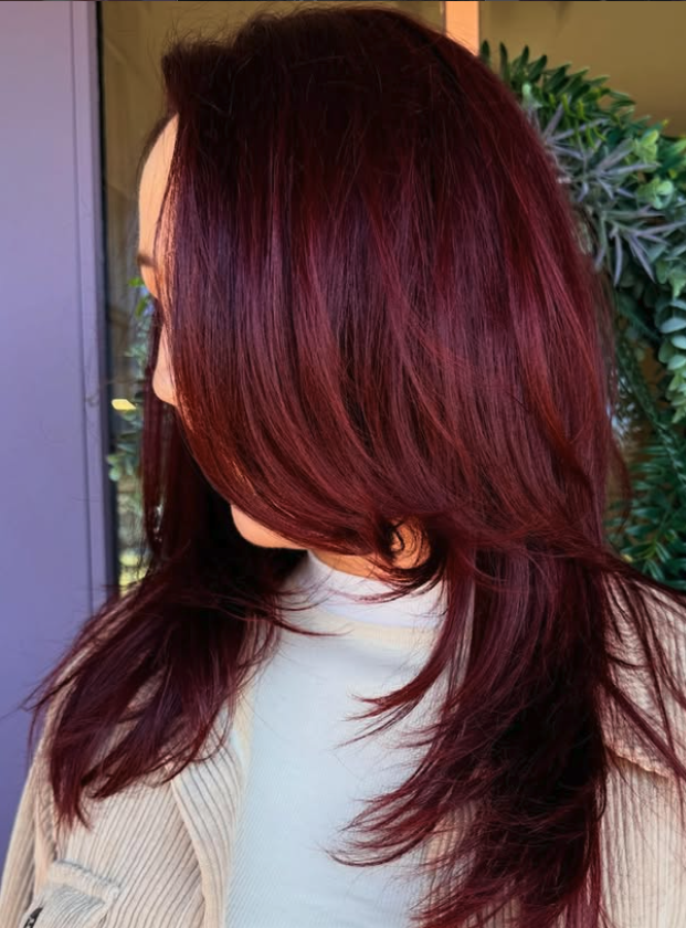 Side profile of a woman with long, layered, deep red hair, standing outdoors near green plants.