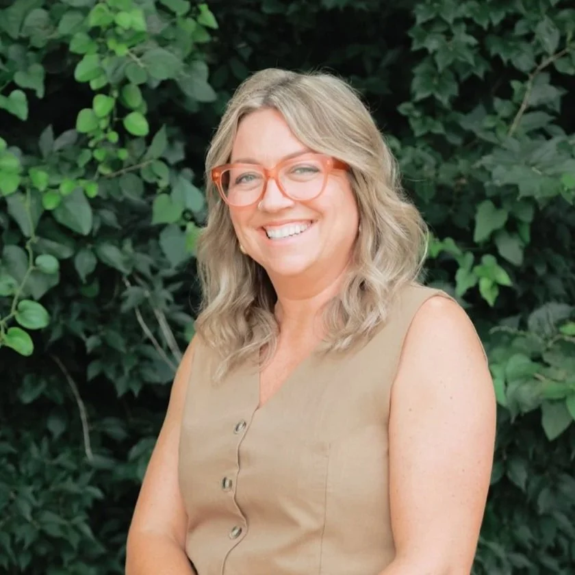 A woman with wavy blonde hair and glasses, smiling outdoors in front of green foliage.