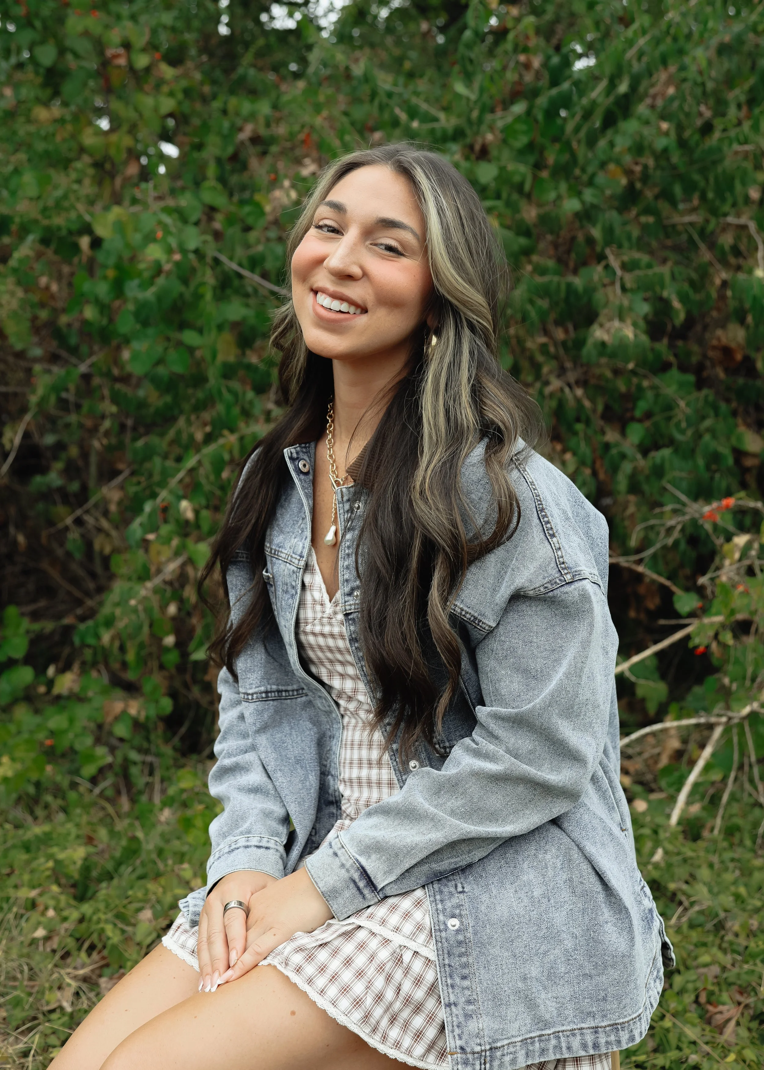 A woman with long dark hair with blonde highlights, smiling, sitting outdoors in front of green bushes, wearing a denim jacket and a checkered dress.